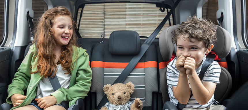 Duas crianças e um peluche sentados nos bancos traseiros de um carro com cinto de segurança